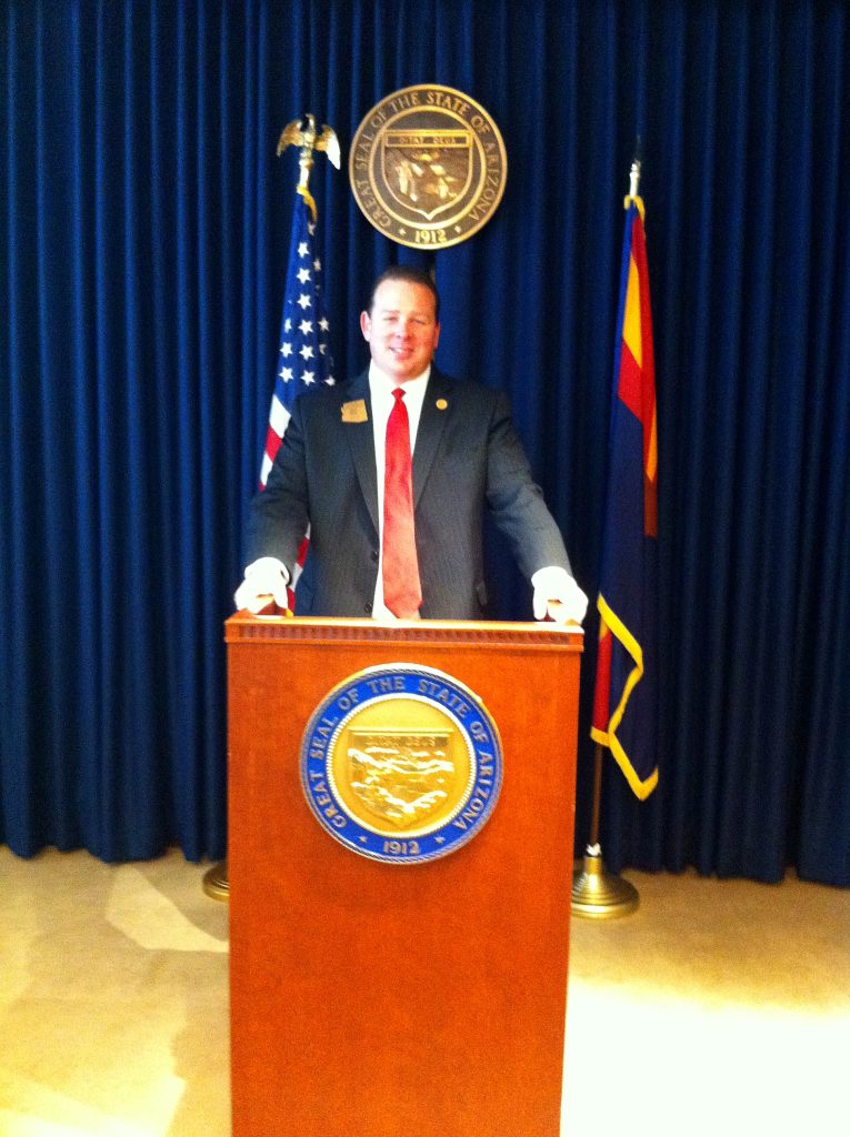 Representative Jeff Dial speaking at a podium at the Arizona State Capitol with the Great Seal of Arizona and the US and Arizona flags