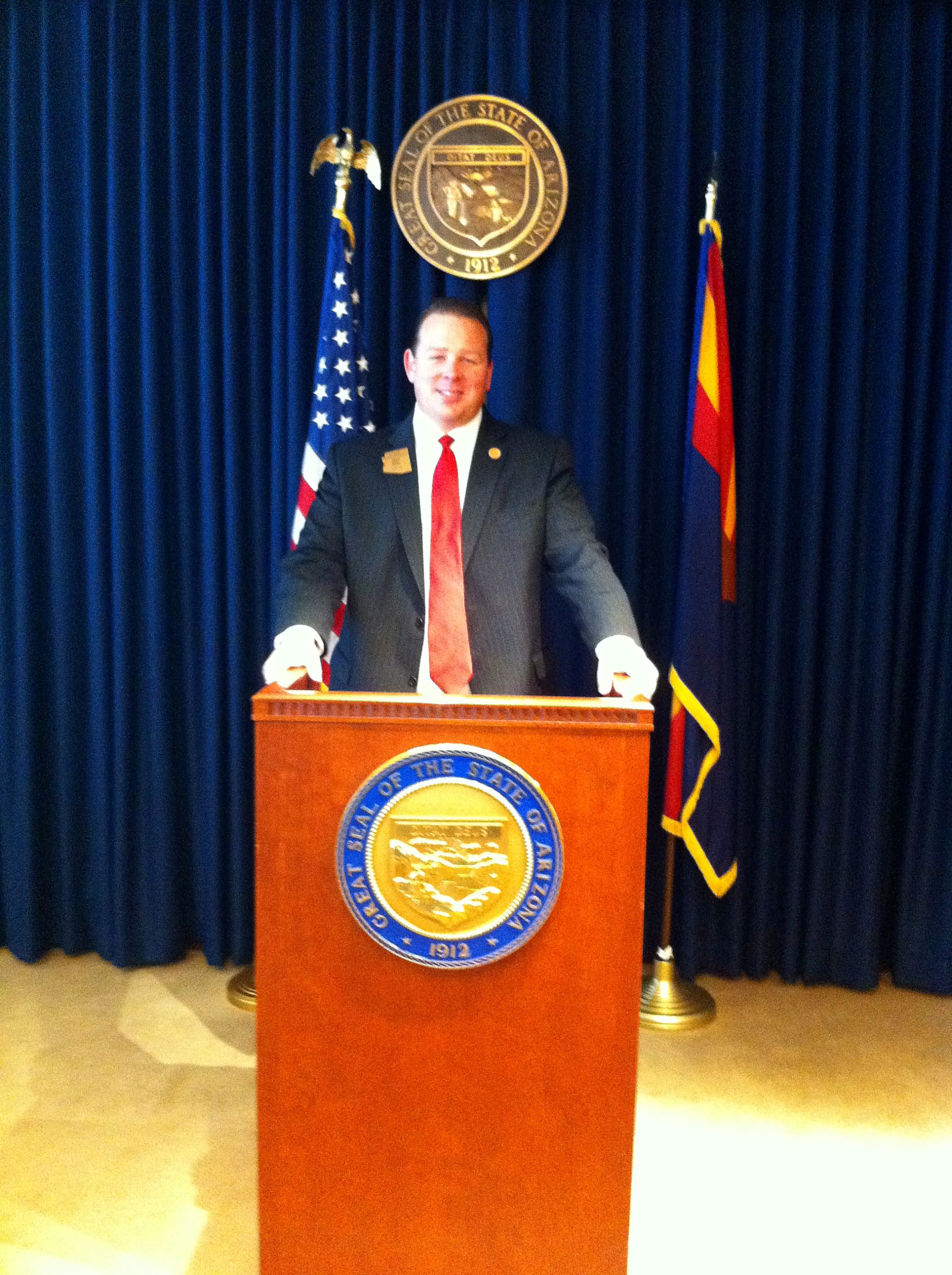 Representative Jeff Dial speaking at a podium at the Arizona State Capitol with the Great Seal of Arizona and the US and Arizona flags